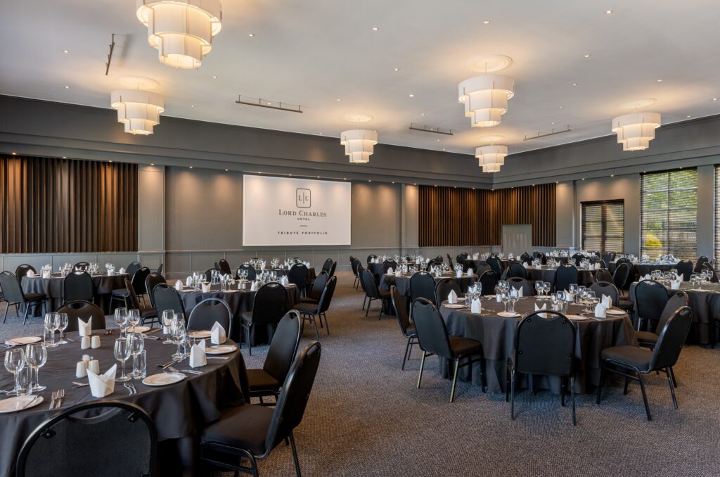 A conference room at The Lord Charles Hotel laid out for a banquet, with round tables, black banqueting chairs, tiered pendant lights and a projection screen at the front.