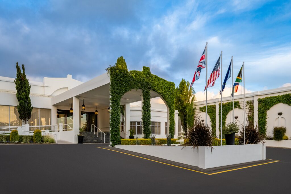 The entrance forecourt of The Lord Charles Hotel: a white portico beside an ivy-covered stone arch, with cypress trees and four flagpoles flying the South African flag.